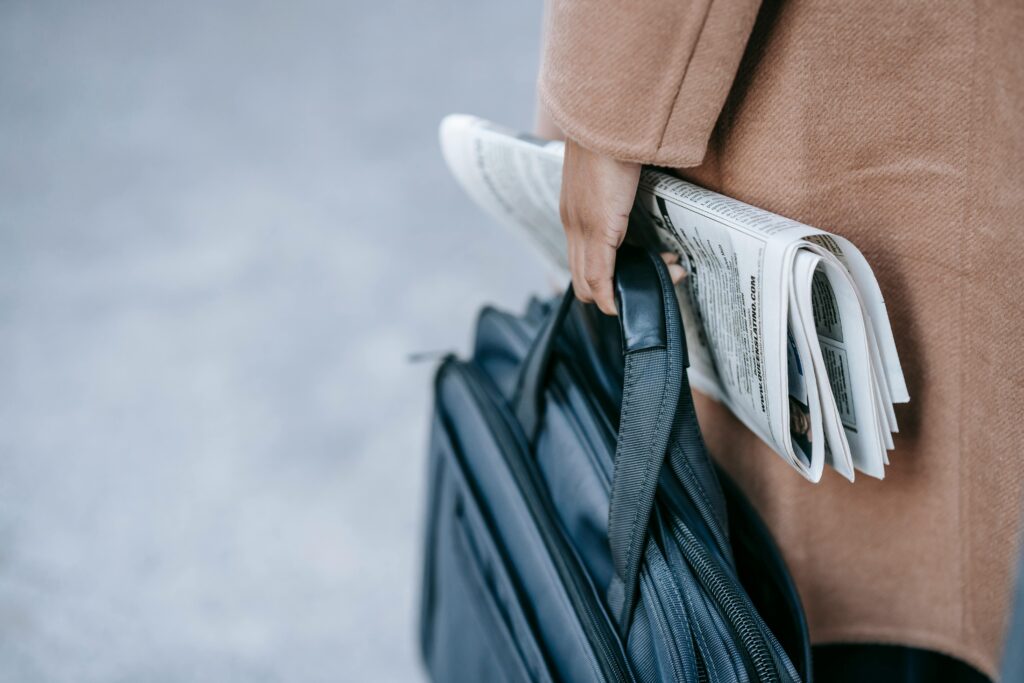 Crop anonymous female entrepreneur in elegant brown coat with laptop in bag and newspaper